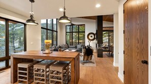 Ampla sala de estar com mesa de jantar de madeira, banquetas de bar e grandes janelas com vista para a floresta.