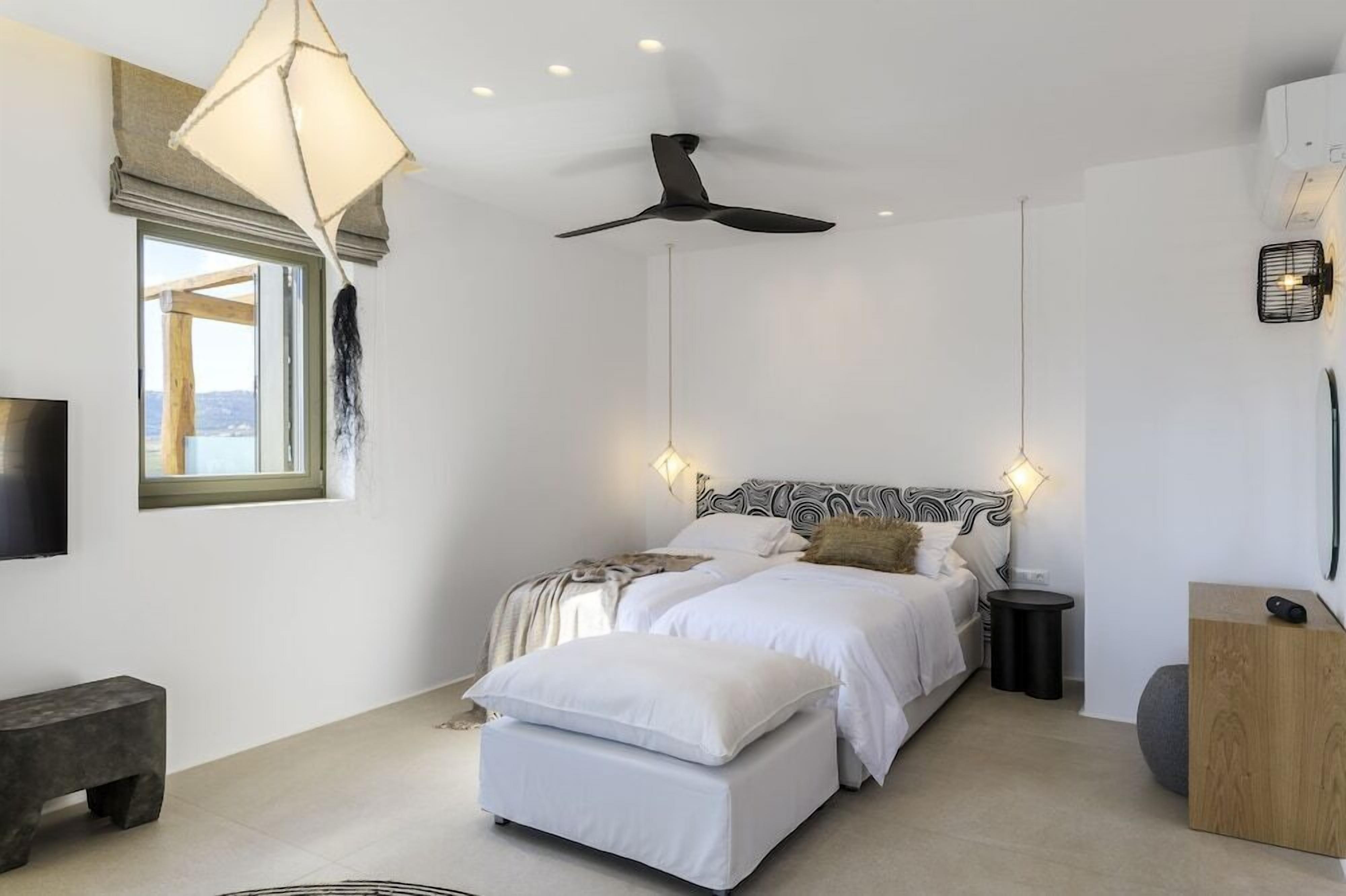 Une chambre à coucher comprenant un lit, un banc, un ventilateur de plafond, une table de chevet et une vue sur l’extérieur.