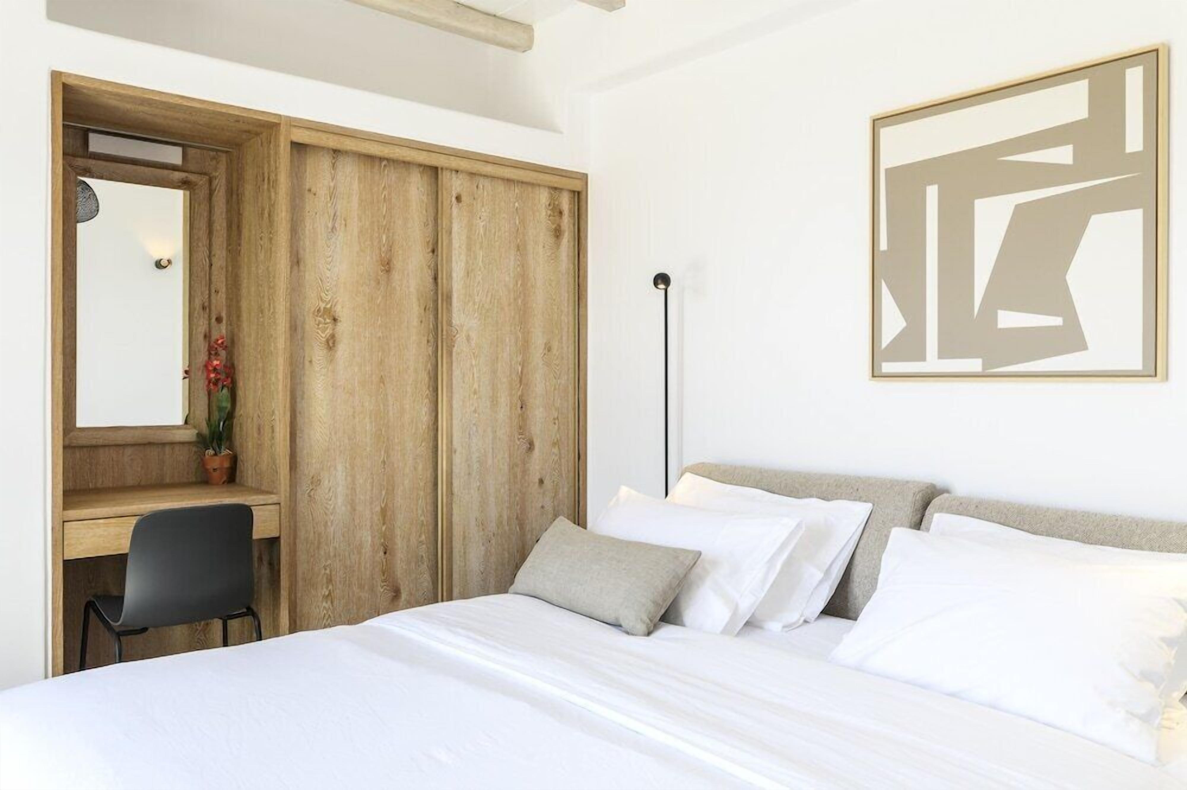 A bedroom with a wooden wardrobe, a desk with a chair, a bed with white bedding, and a floor lamp.