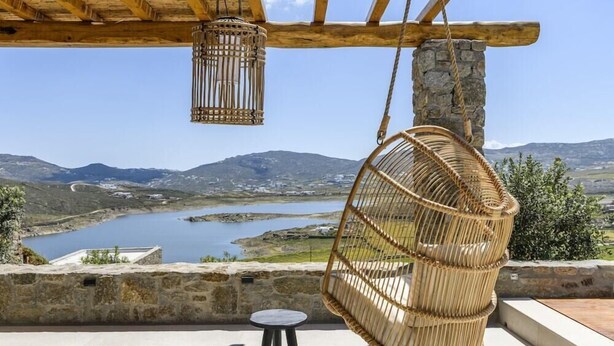 A wicker chair hanging from a wooden beam, a small round table, and a stone wall overlooking a valley with a lake and mountains. - Nasta Intentional Living Mykonos (Mykonos)