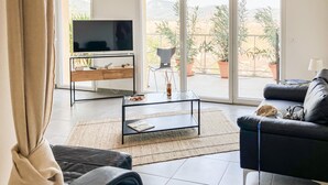 A modern living room with a TV, sofa, coffee table, and a view of a balcony with potted plants. - Appt proche plage et St-Tropez (Grimaud)