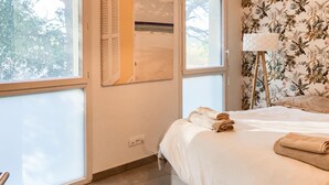 A bedroom with a bed, a window, a lamp, and a wall with floral wallpaper. - Appt proche plage et St-Tropez (Grimaud)