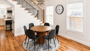 Un coin repas avec une table ronde en bois, six chaises, un lustre, un escalier et une cuisine en arrière-plan.