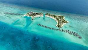 Vista aérea de una isla resort con bungalows sobre el agua, un edificio central y una playa.