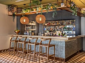 Uma área de bar com chão xadrez, cadeiras de madeira, plantas pendentes e prateleiras com garrafas de bebidas.