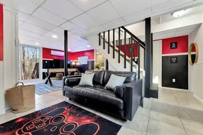 A modern living room with a black leather sofa, a red and black wall, a staircase, and a gaming area in the background. - Modern 5BR GameRoom, HotTub, FirePit + Lake Access (East Stroudsburg)
