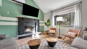 Una sala de estar con chimenea, dos sillones, una mesa de centro, una lámpara de pie y un ventanal con cortinas.