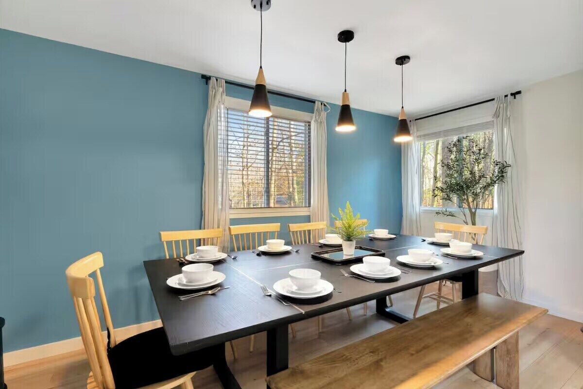A dining area with a long table set for four, wooden chairs, pendant lights, and a window with curtains.