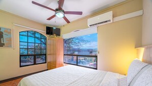 Un dormitorio con cama, ventilador de techo, equipo de aire acondicionado, televisor y ventanas amplias con vistas.