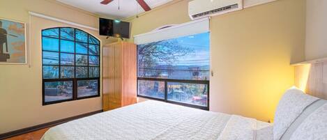 Un dormitorio con cama, ventilador de techo, equipo de aire acondicionado, televisor y ventanas amplias con vistas.