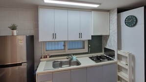 Une cuisine de taille réduite avec des meubles blancs, un réfrigérateur en acier inoxydable, une cuisinière à gaz et une horloge murale.