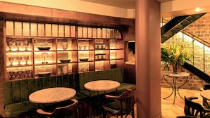 A restaurant with a green cushioned bench seating area, multiple round tables, and a brick wall with shelves displaying glassware and plates. - 5Q House Chapinero (Bogotá)
