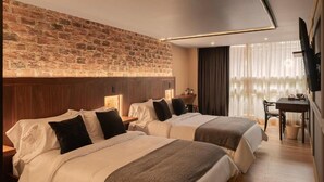 A hotel room with two beds, a stone wall, a flat-screen TV, a dining table, and a window with curtains. - 5Q House Chapinero (Bogotá)