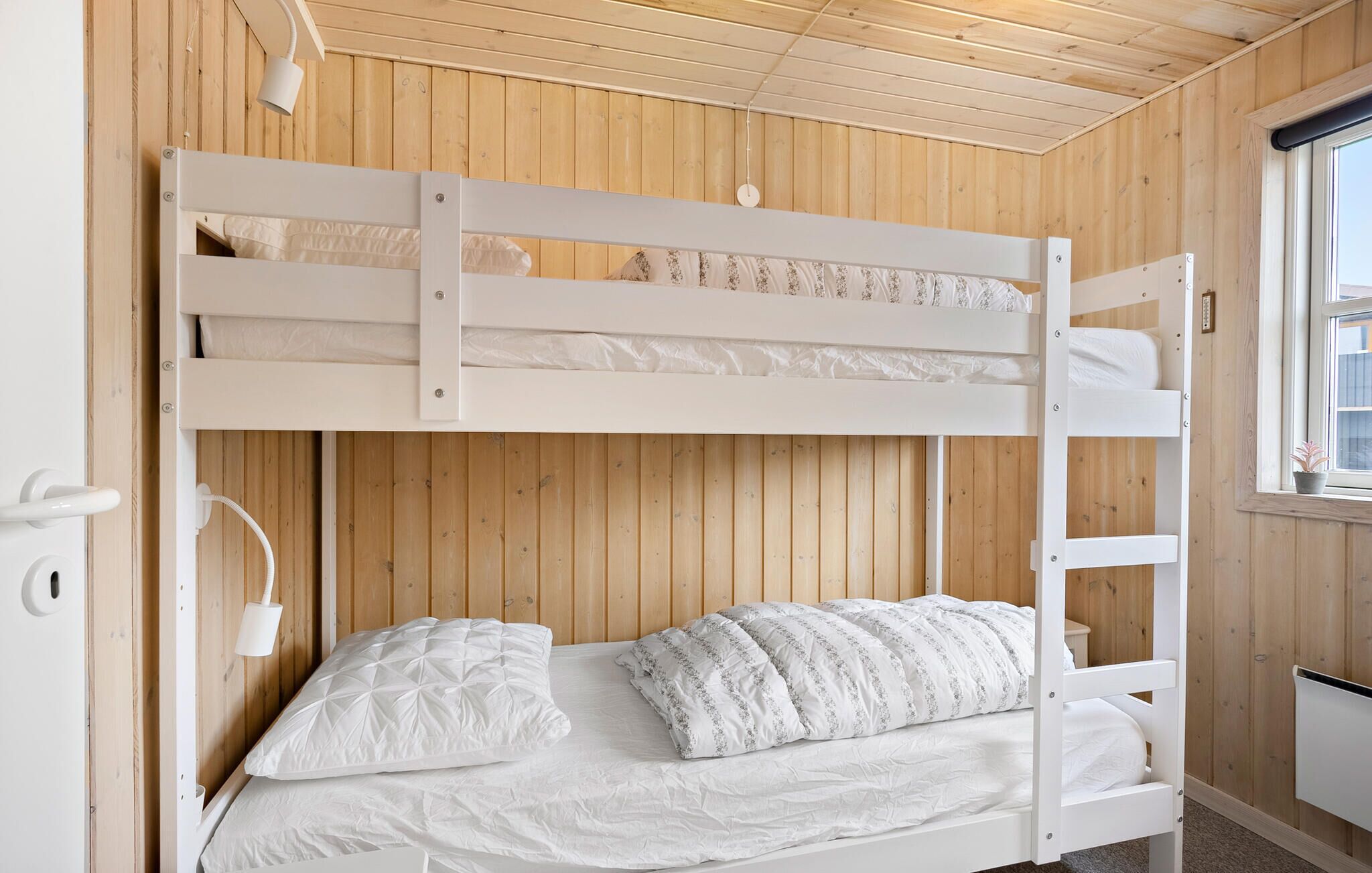 Een stapelbed met witte beddengoed, een houten wand, een raam en een kleine potplant.