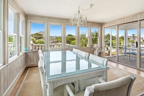 A spacious room with a large glass table and white chairs, large windows with a view of greenery, and a chandelier. - Spacious 7BR coastal retreat, pet-friendly, ocean views, large yards, perfect for families & groups (North Myrtle Beach)