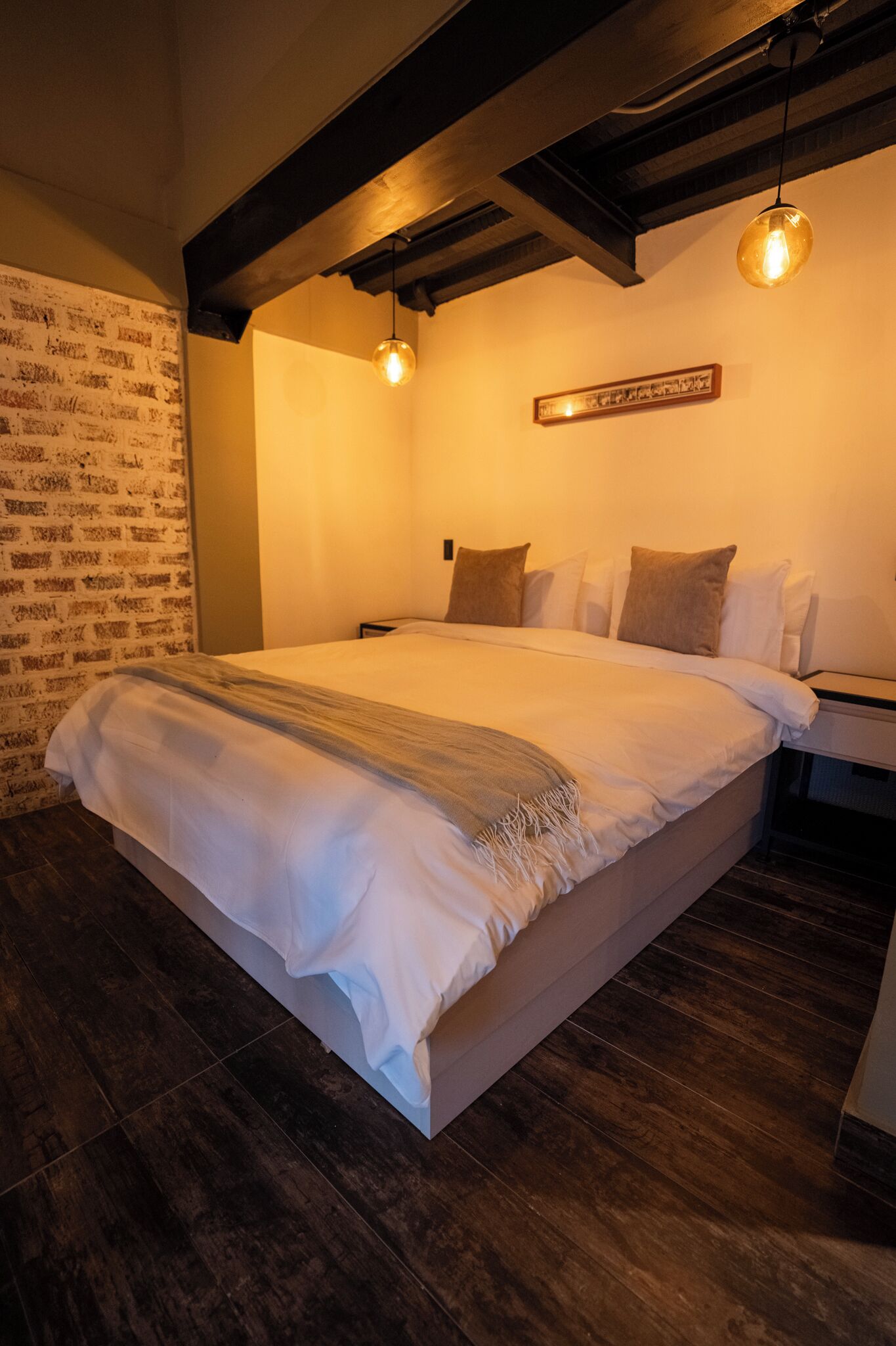 Una cama bien tendida con una colcha, pared con patrón de ladrillos y una viga de madera en el techo.