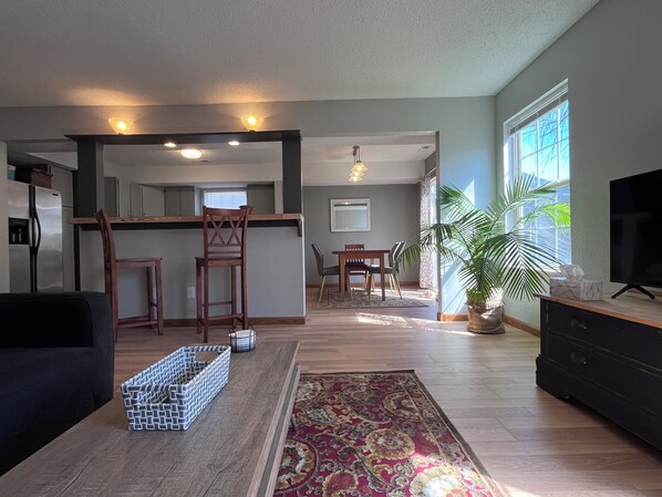 A living room with a sofa, a coffee table, a TV, a dining area with chairs and a table, a kitchen area with a fridge, and a large window with a plant.