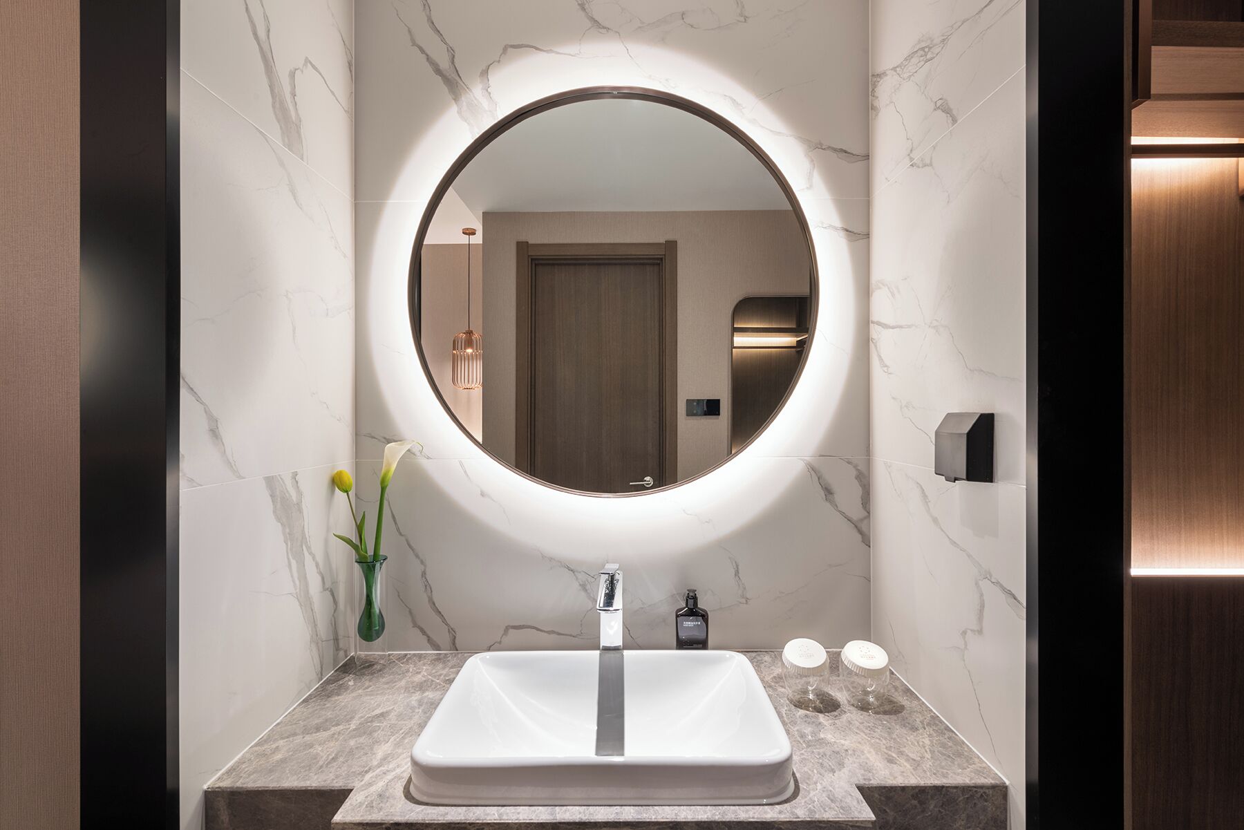 A bathroom with a round mirror, a white sink, marble walls, and a wooden door.