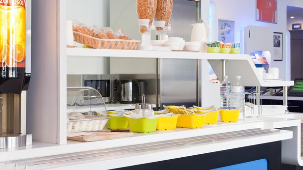 A breakfast bar with cereal, coffee, and water dispensers, and a basket of bread. - Ibis Budget Berlin Ost (Berlin)