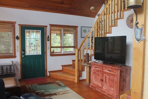 A living room with a staircase, wooden floors, a green door, a television on a red cabinet, and a clock on the wall. - 2 bedroom 2 bath cozy cabin on 40 acres in Fairplay. (Fairplay)