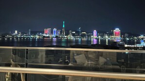 A nighttime cityscape with illuminated buildings across a body of water, viewed from a balcony with a glass railing. - Cozy studio for couple and small family in Sapphire Ha Long (Ha Long)