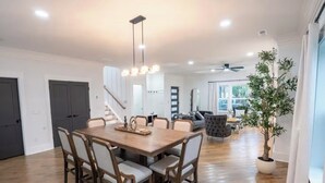 Uma ampla sala de jantar com uma mesa e cadeiras de madeira, uma escada, uma sala de estar com um sofá e um vaso de planta, e um ventilador de teto.