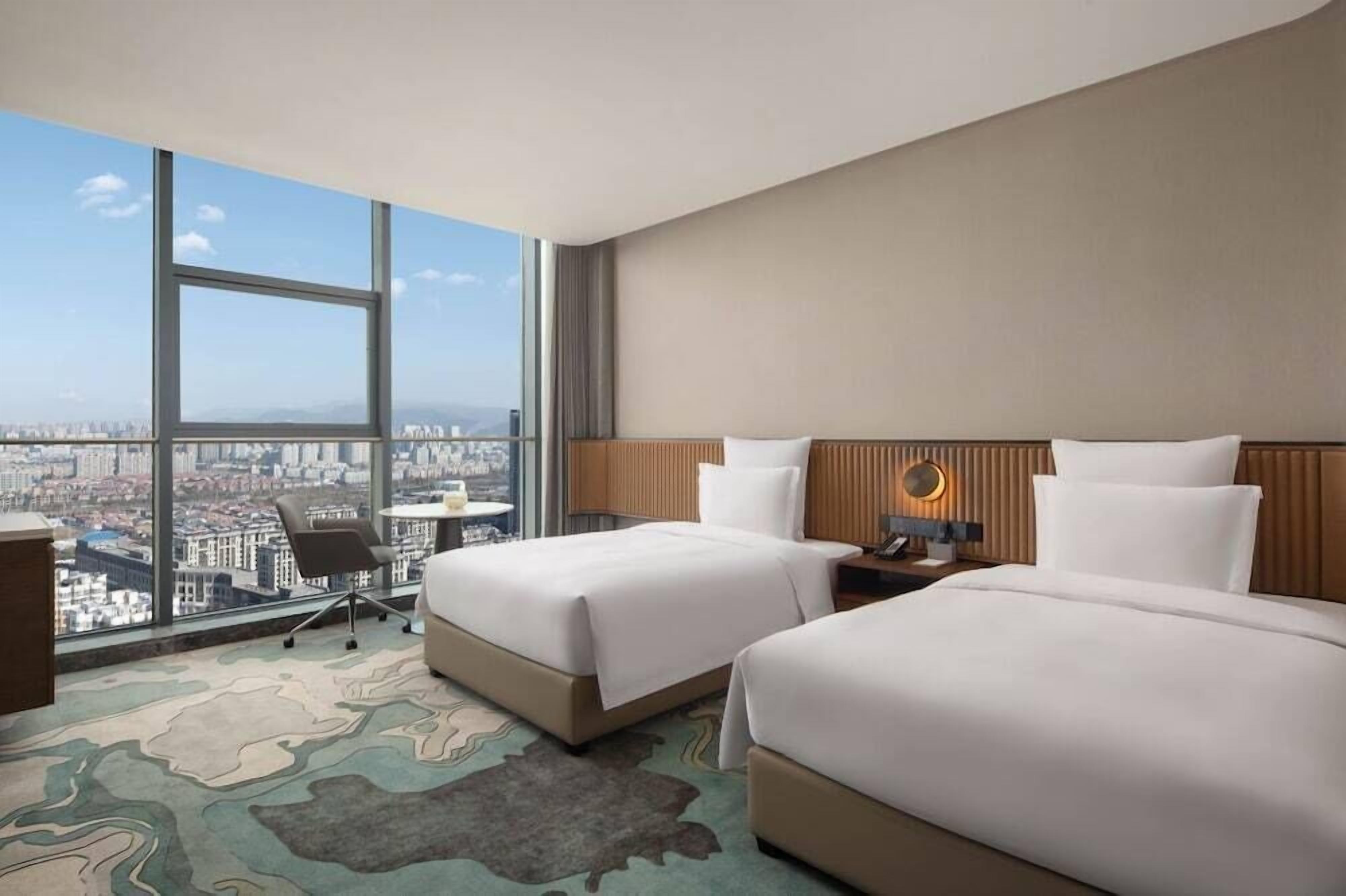 A hotel room with two beds, a desk, and a chair. There is a large window offering a city view.