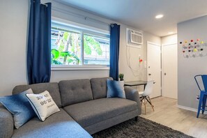 Una sala moderna con un sofá seccional gris, una mesita auxiliar, una silla blanca y una ventana con cortinas azules.