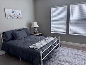 Une chambre à coucher avec un lit, une table de chevet avec une lampe, deux fenêtres équipées de stores et un tableau accroché au mur.