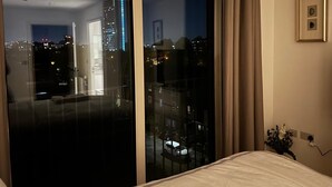 Une pièce dotée d’une grande porte-fenêtre coulissante donnant sur un balcon extérieur, d’un lit recouvert d’une couverture blanche, d’un tableau encadré au mur et d’une prise électrique avec un câble.
