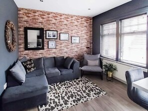 Un salon avec un mur en briques, un canapé gris, un tapis à motifs et une fenêtre avec des stores.