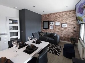 A modern living room with a dining area, a grey sofa, and a brick wall with framed pictures. - Modern Apartment - Sleeps 4 - Centrally located (Lanark)