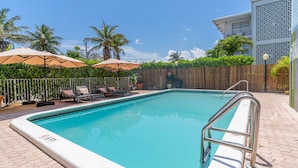 Una piscina rectangular con barandilla de seguridad metálica, rodeada de sillas de descanso y sombrillas, con cerca de madera y palmeras al fondo.