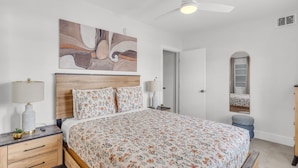 Un dormitorio con cabecera de madera, ropa de cama floral, una lámpara de noche, un ventilador de techo, una puerta y un espejo.