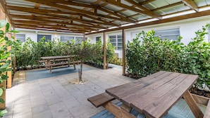 Un área de patio techada con una mesa y bancos de madera, rodeada de abundante follaje.