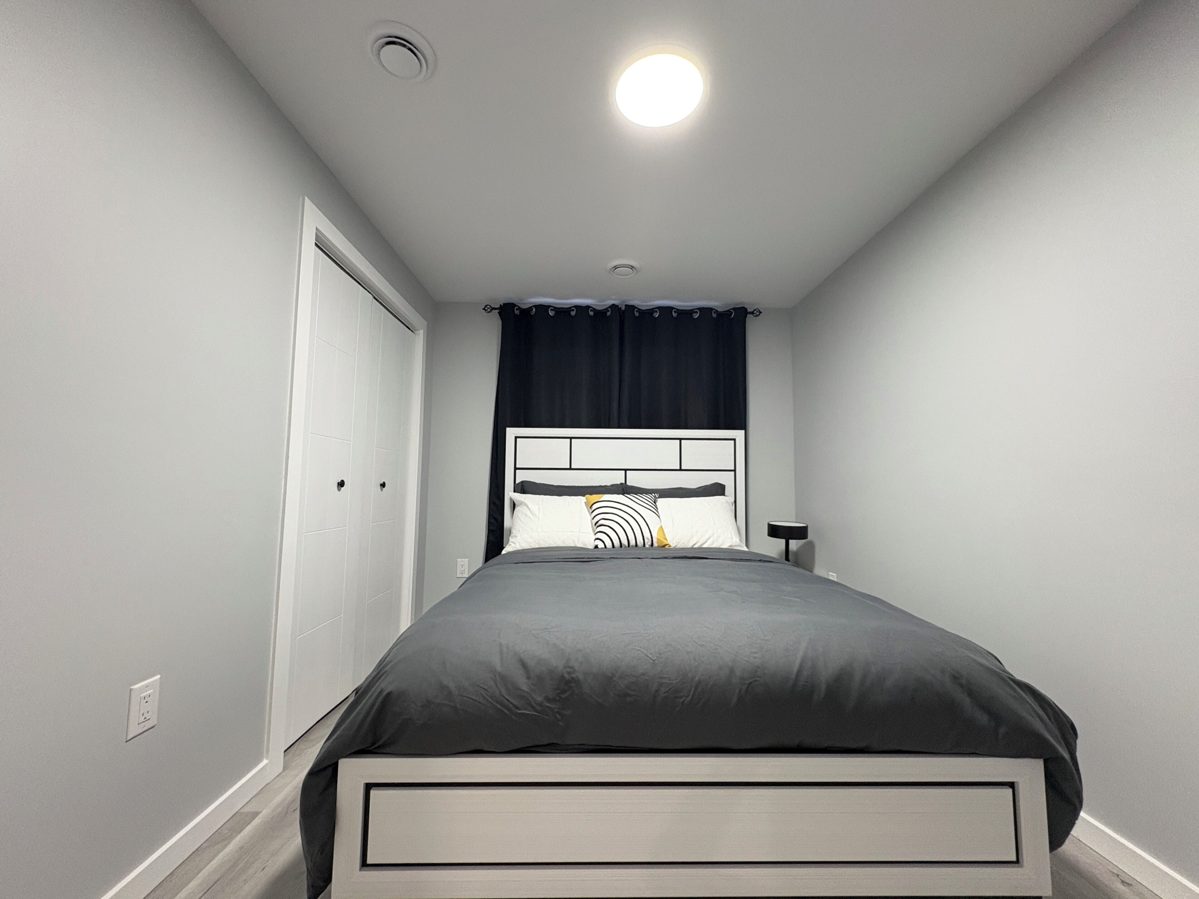 Een slaapkamer met een bed, nachtkastje, plafondlamp en een deur.