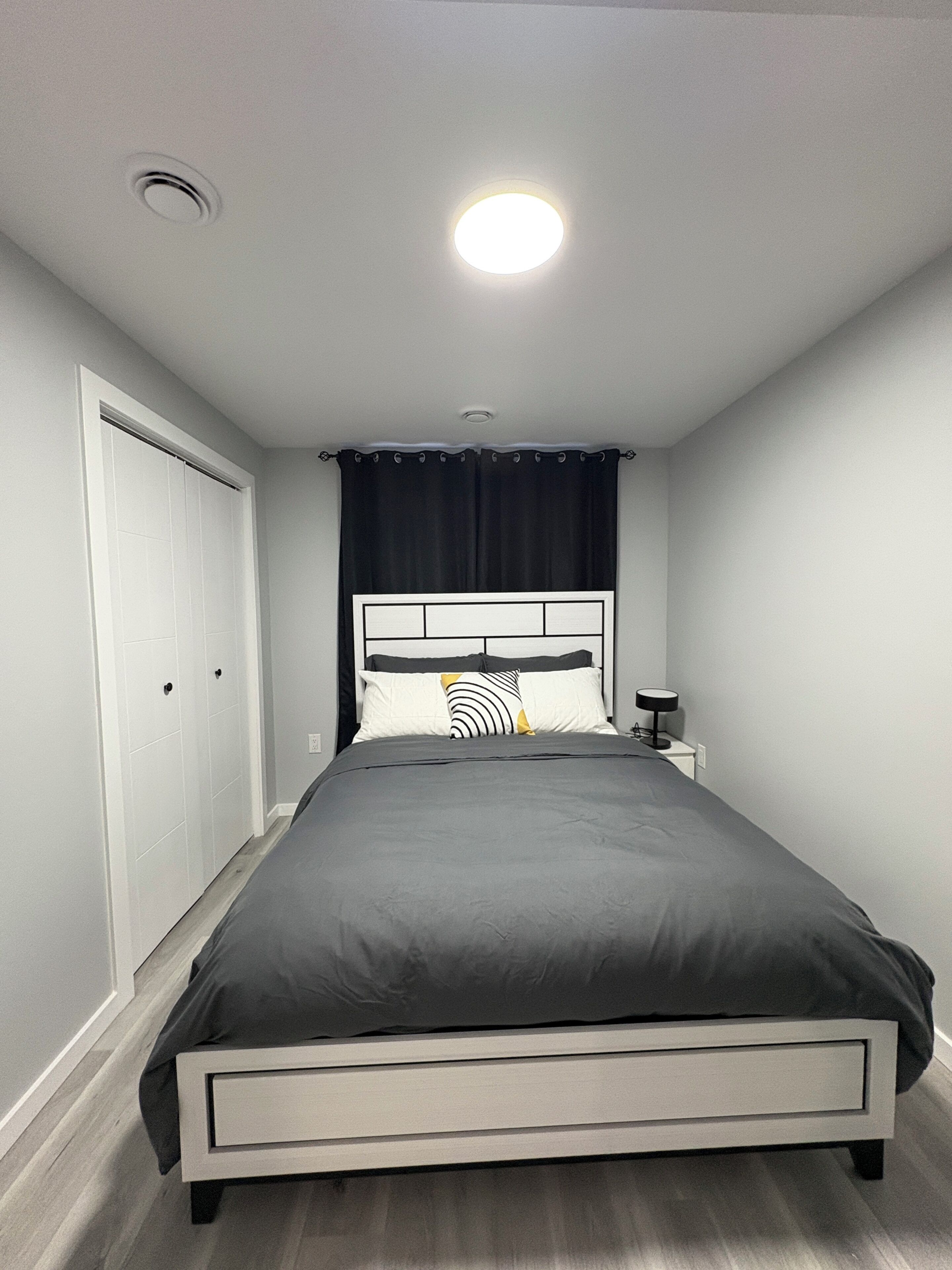 Un dormitorio con una cama, una mesita de noche, una lámpara, una luz de techo y una puerta.