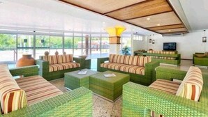 Un hall d’entrée spacieux, doté de canapés confortables recouverts d’un tissu vert, d’une grande fenêtre donnant sur l’extérieur et d’un téléviseur fixé au mur.