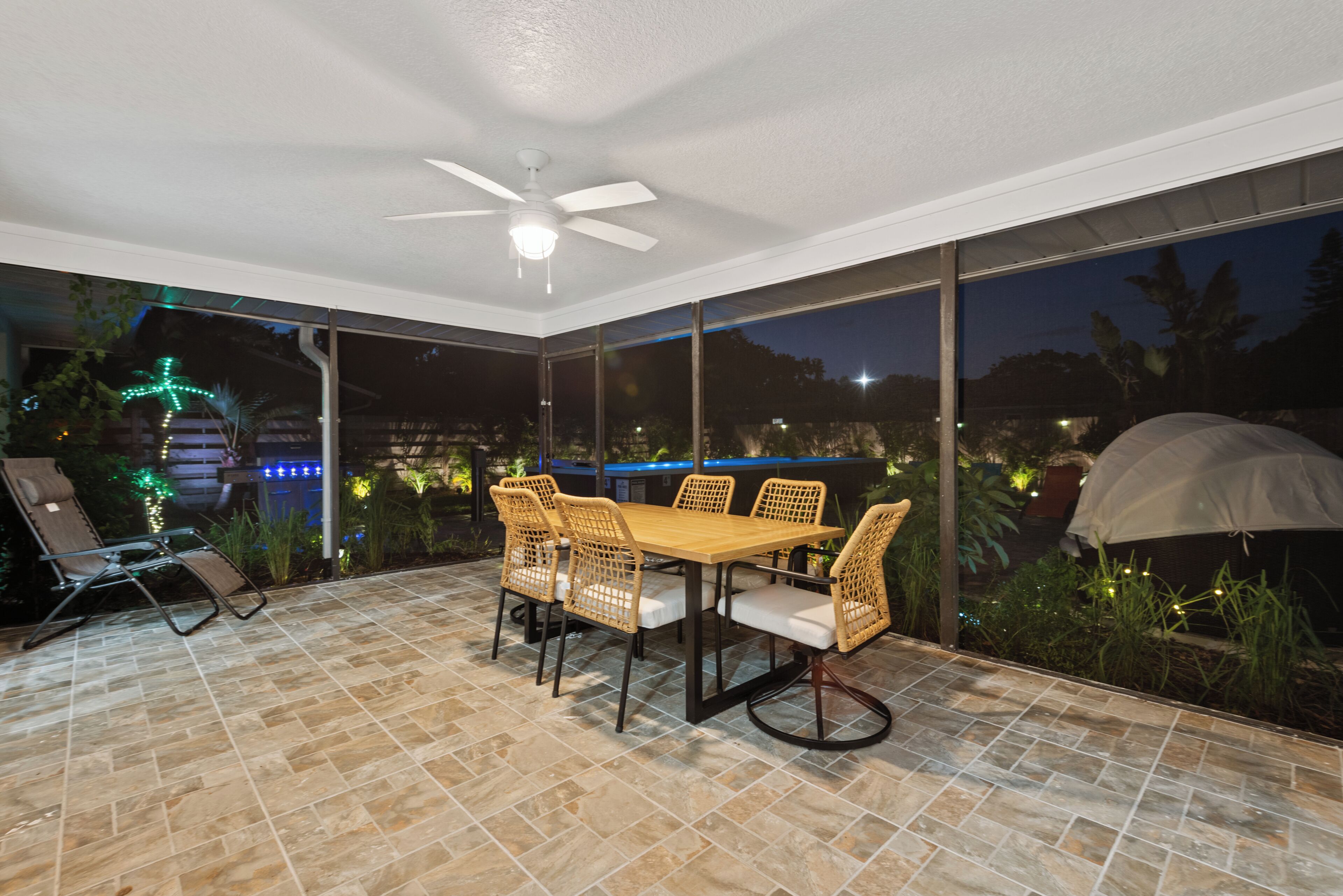 An outdoor patio with a dining table and chairs, a ceiling fan, and large windows offering a view of the surrounding area.