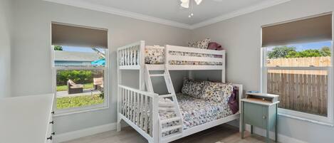 A bedroom with a bunk bed, a ceiling fan, a window with a view of the outdoors, and a small bedside table.