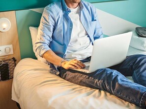 A person using a laptop on a bed in a hotel room with a wooden headboard and a wall socket. - Ibis Budget Les Sables Olonne Sur Mer (Les Sables-d'Olonne)