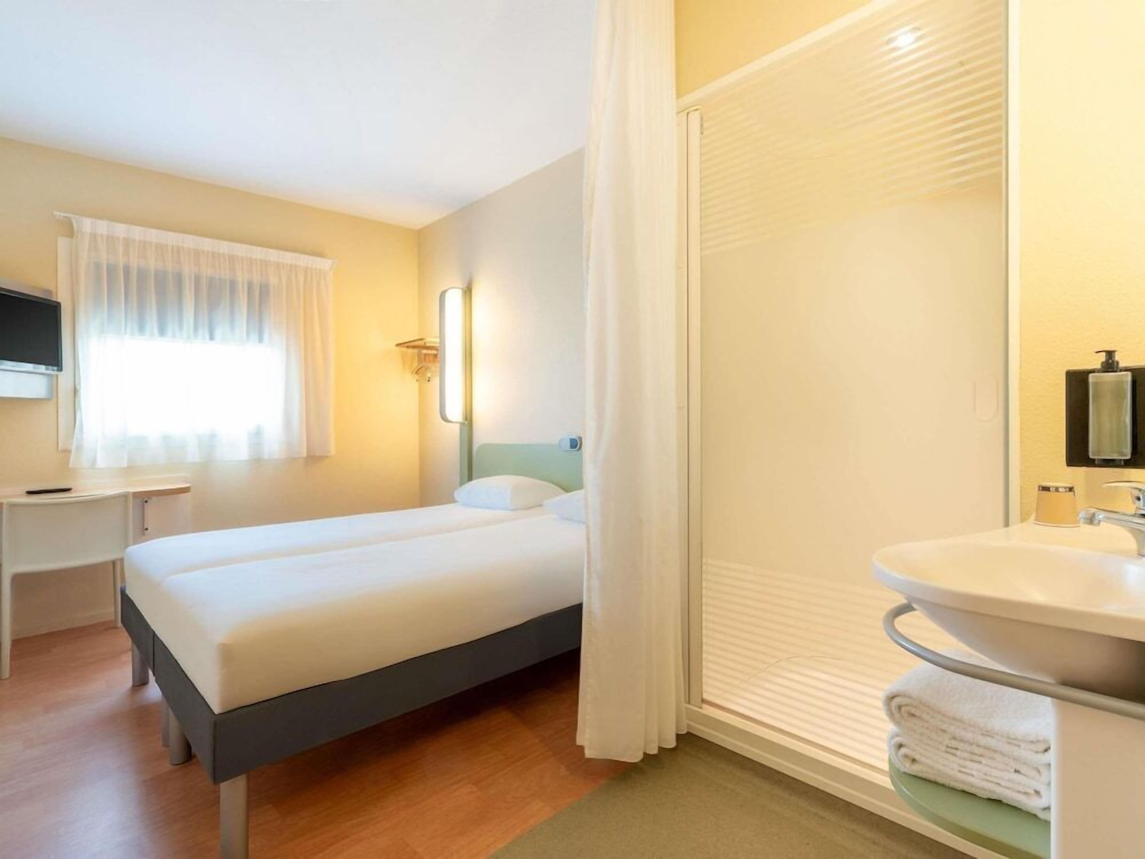 Ein Hotelzimmer mit Bett, Schreibtisch und einem Badezimmer mit Waschbecken und Handtuchhalter.