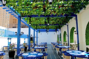 An outdoor cafe with tables and chairs, a blue railing, and greenery overhead. - Planet Hollywood Thane City (Thane)