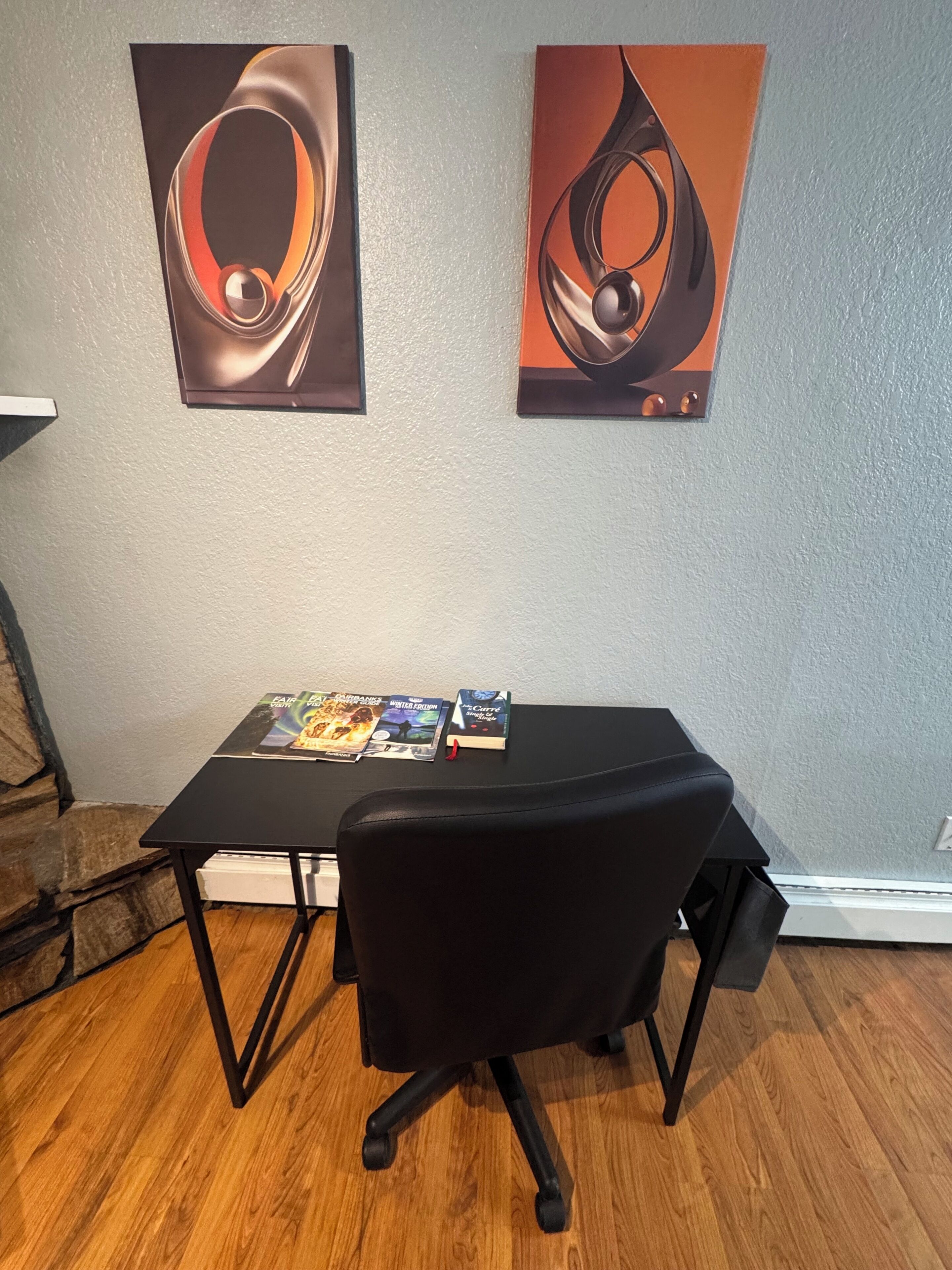 A room with a black office chair, a table with magazines, and two abstract paintings on the wall.