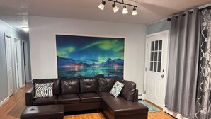 A modern living room with a sectional sofa, a wooden floor, a large painting of a night sky, and a door with curtains. - Central Fairbanks 3BR/2BA,  King Bed,  Sleeps 6, Free Laundry (Fairbanks)
