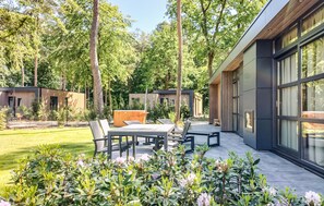 Une terrasse extĂ©rieure moderne, dotĂ©e dâune table et de chaises, et entourĂ©e dâune vĂ©gĂ©tation luxuriante et de grands arbres.