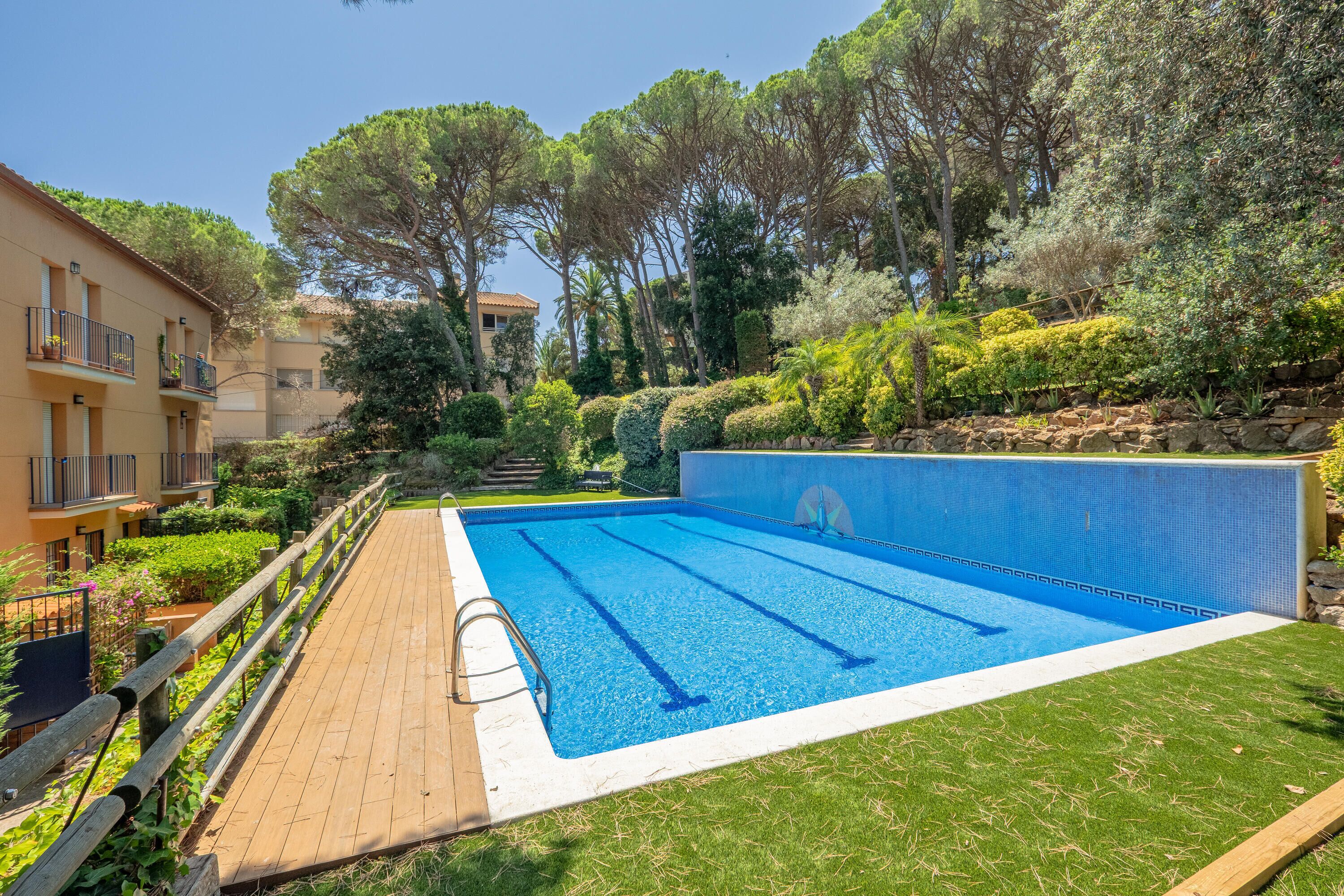 A swimming pool with a wooden deck, surrounded by greenery and a building with balconies.