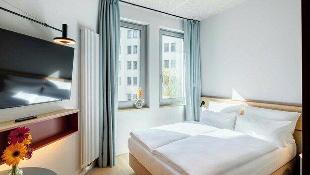 A hotel room with a bed, a TV, a small table with flowers, and a window with curtains.