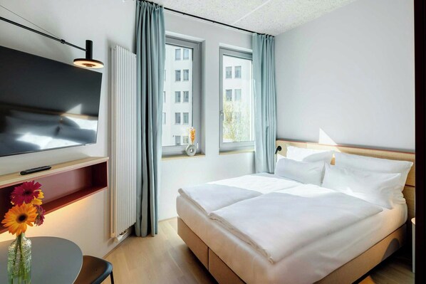 Ein Hotelzimmer mit einem Bett, einem Fernseher, einem kleinen Tisch mit Blumen und einem Fenster mit Vorhängen.