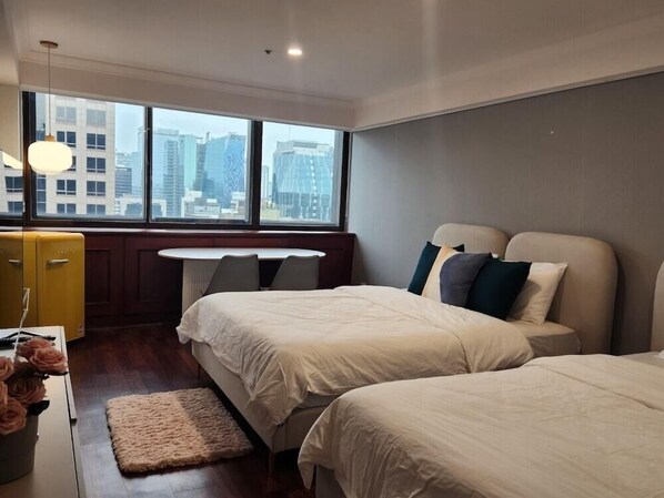 A modern hotel room with two beds, a small table, a yellow cabinet, and a view of city buildings.